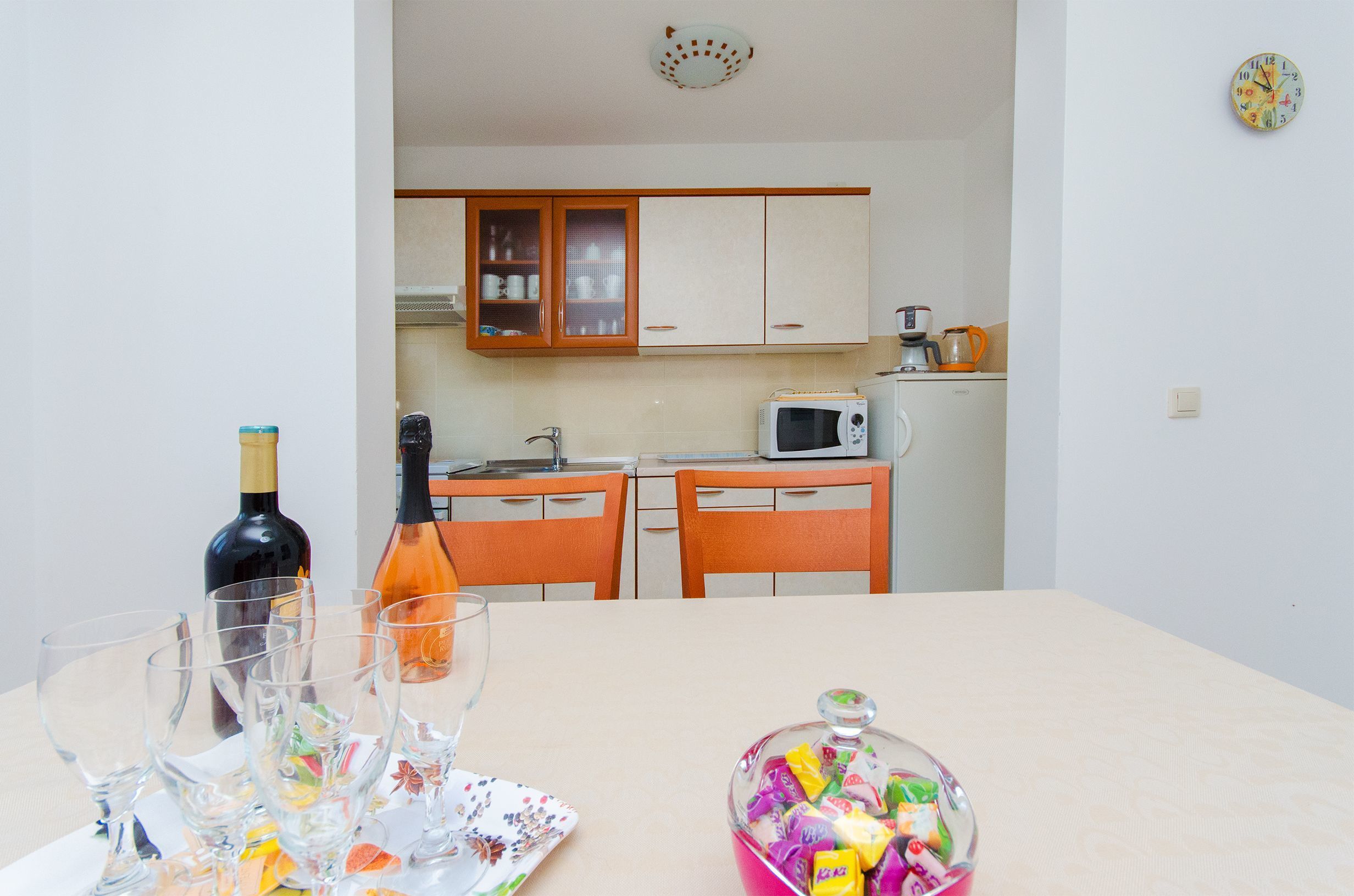A dining table with wine glasses, a bottle, and a glass pitcher. A microwave and kettle are on the kitchen counter.