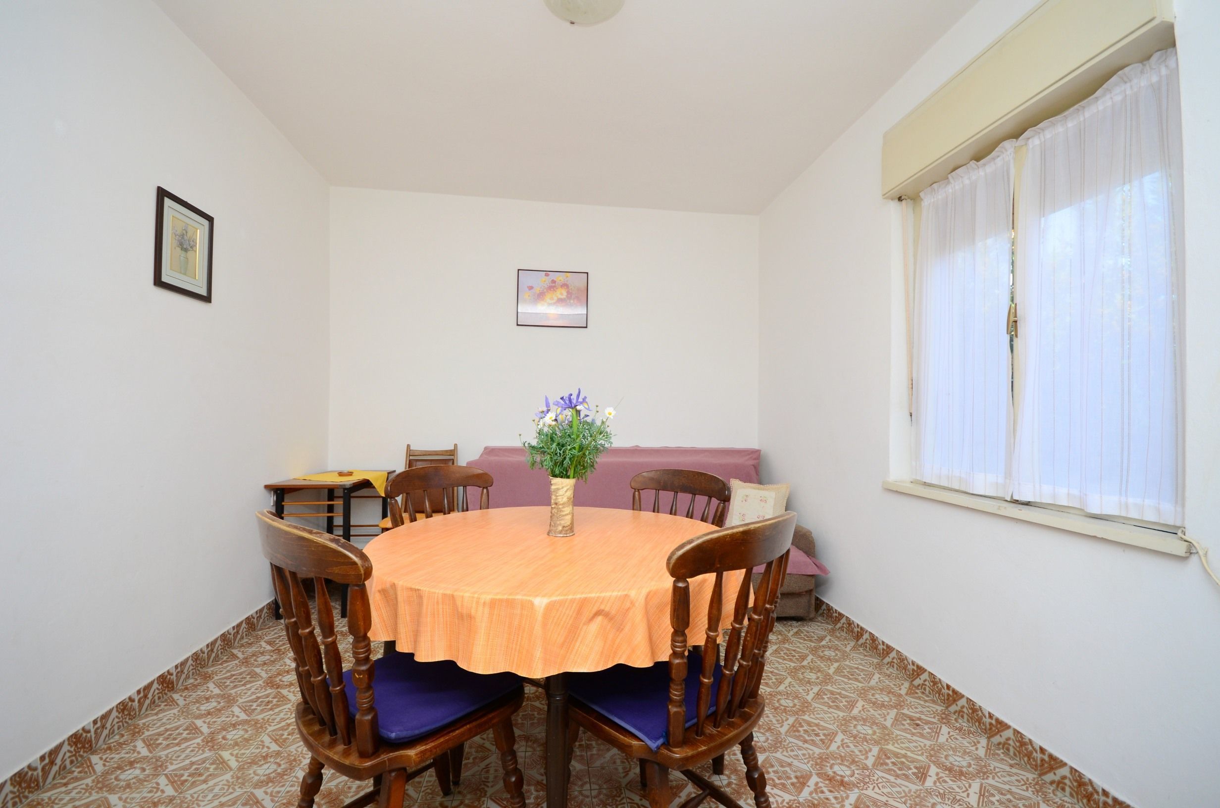 A dining area with a round table, chairs, a vase with flowers, a sofa, and a window with curtains.