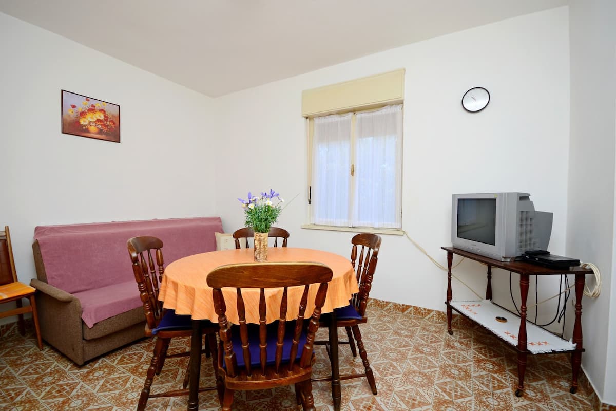 Un área de comedor con una mesa redonda, sillas, un sofá, un televisor sobre un mueble bajo y un reloj en la pared.