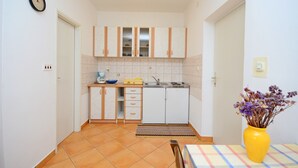 Uma cozinha compacta com armários brancos, uma pia e uma bancada pequena. Há um relógio na parede e um vaso com flores na mesa.