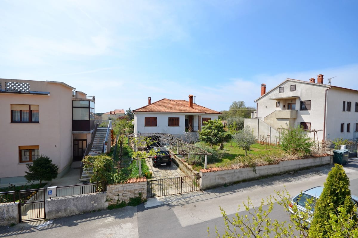 Un barrio residencial con una casa blanca de techo de tejas rojas, una escalera que lleva a un balcón y un pequeño jardín.