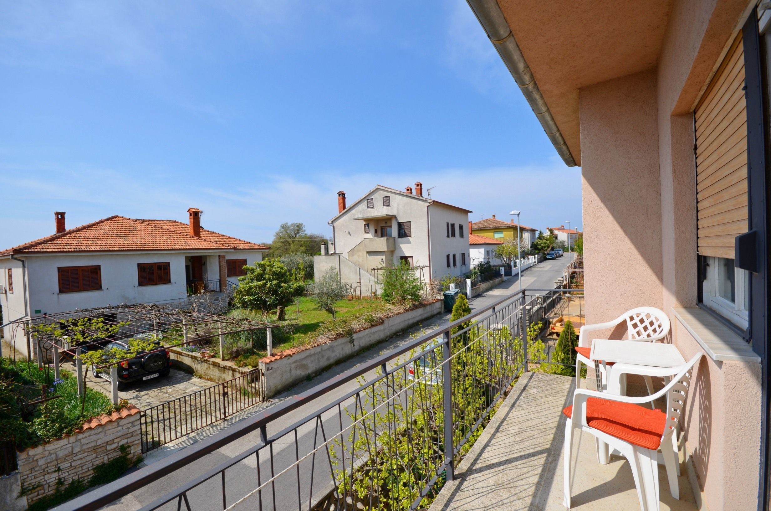 A balcony with a view of a residential street, a parked car, and a white chair.
