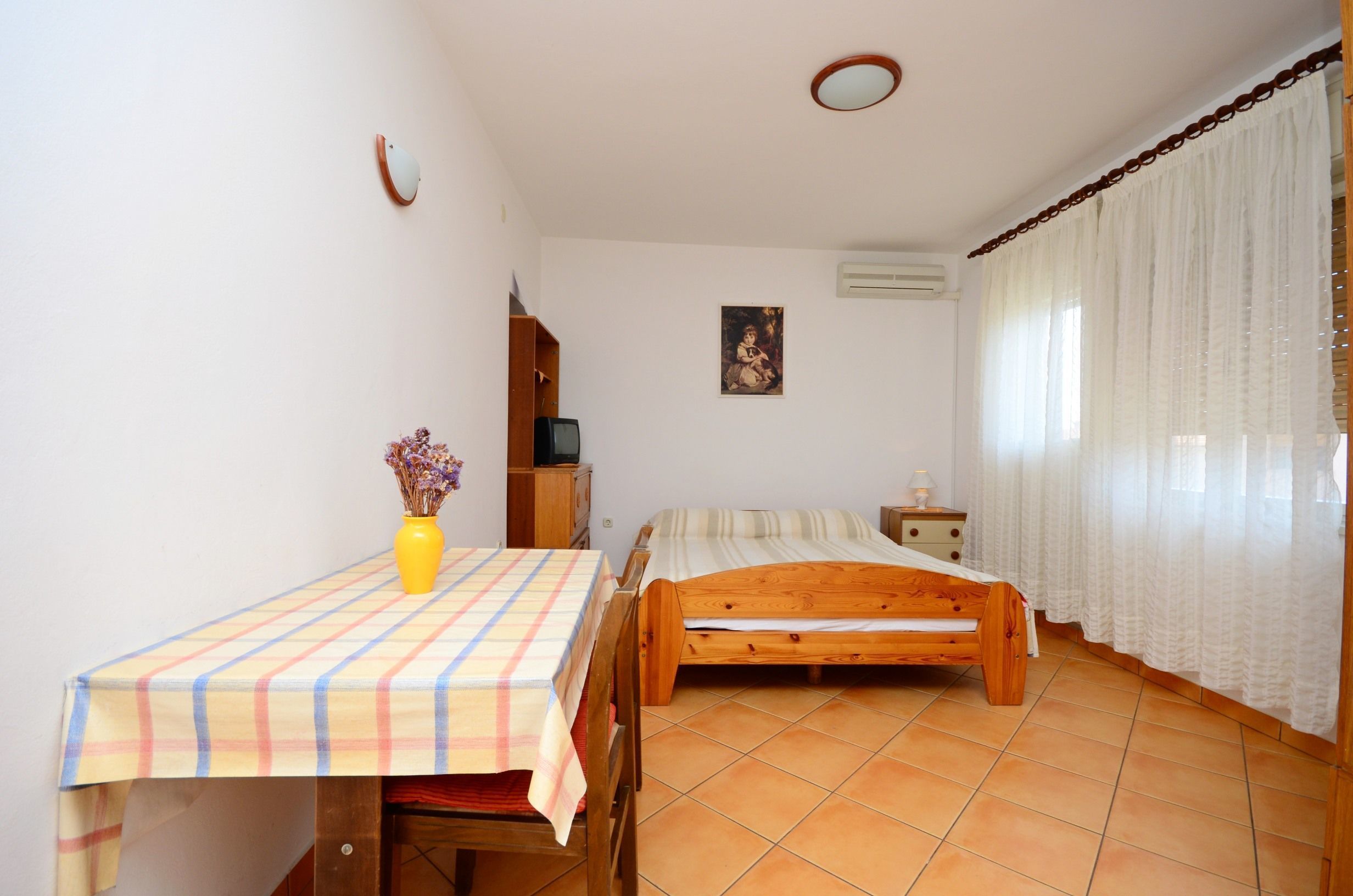 Une chambre à coucher avec un lit, une table recouverte d’une nappe à carreaux, une table de chevet en bois, une petite armoire et un climatiseur fixé au mur.