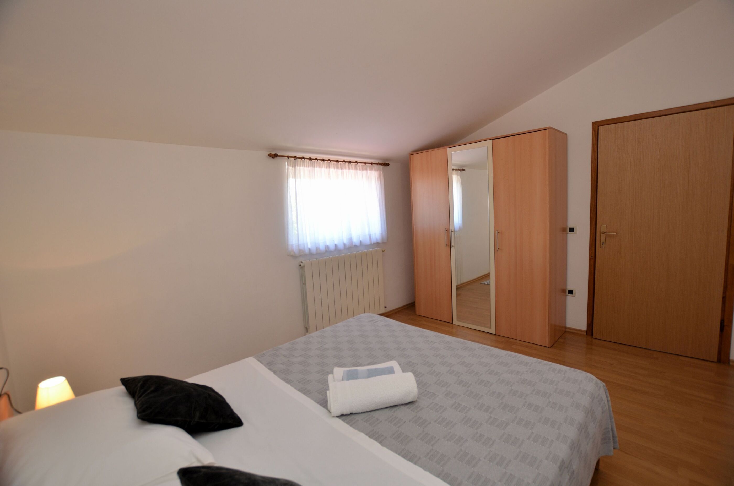 Un dormitorio con cama, ventana con cortinas, radiador, armario y puerta.