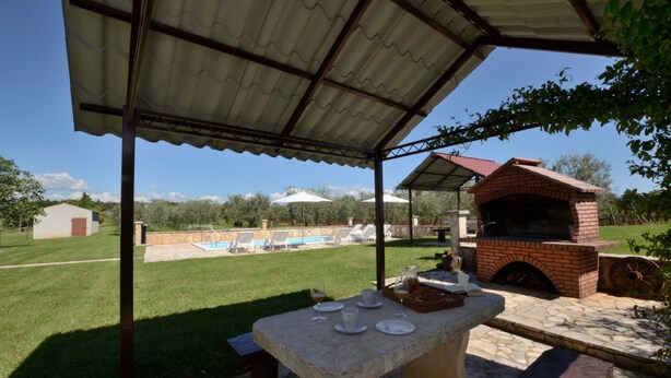 Un espacio de estar al aire libre con techo, una mesa de piedra, un horno de ladrillo y una piscina al fondo.