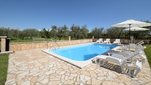 Una piscina con baldosas de piedra, rodeada de sillas de lounge blancas y sombrillas, con un muro de piedra y vegetación de fondo.