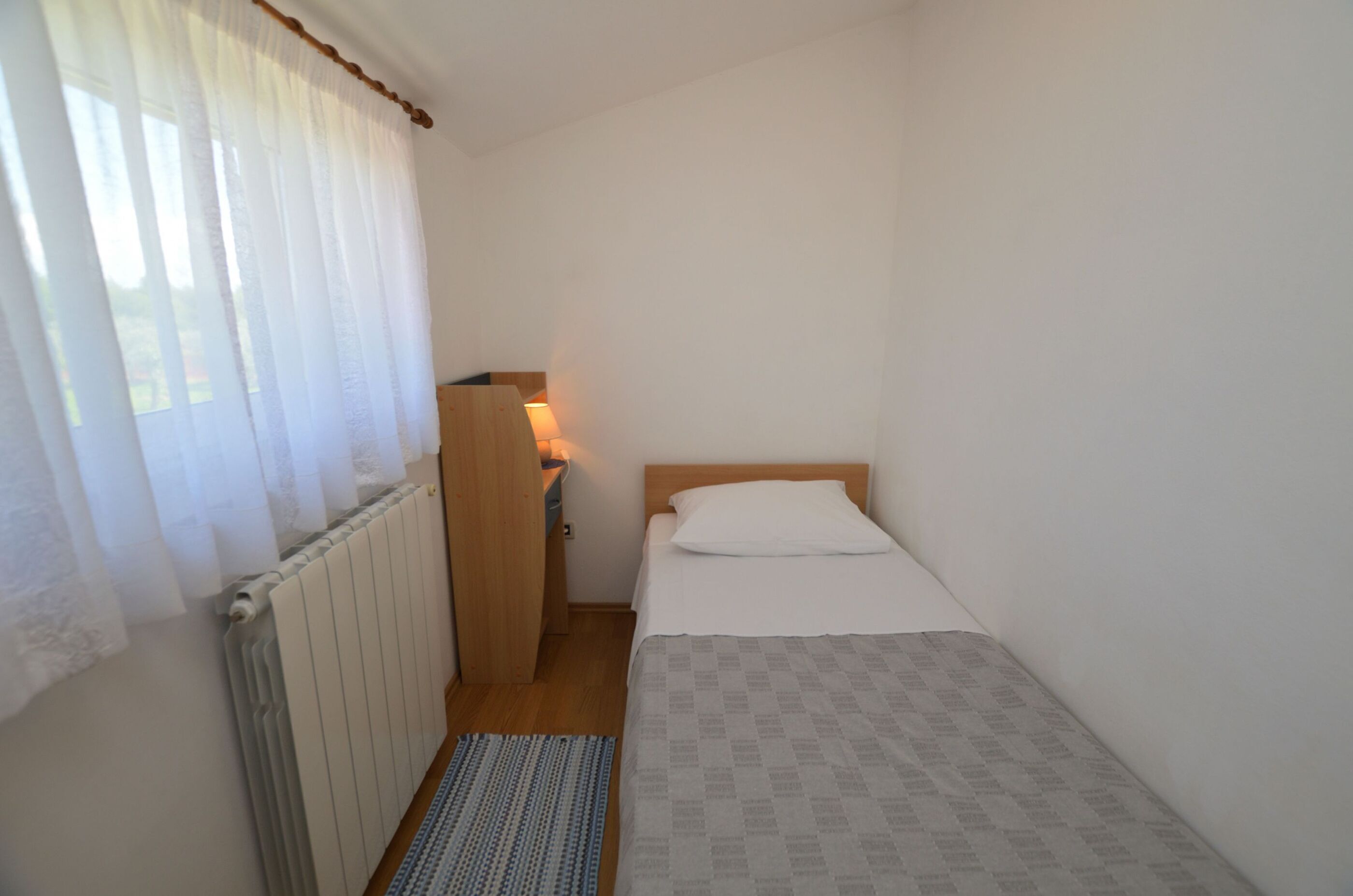 Una habitación pequeña y limpia con una cama individual, una mesita de noche de madera, un radiador y una ventana con cortinas translúcidas.