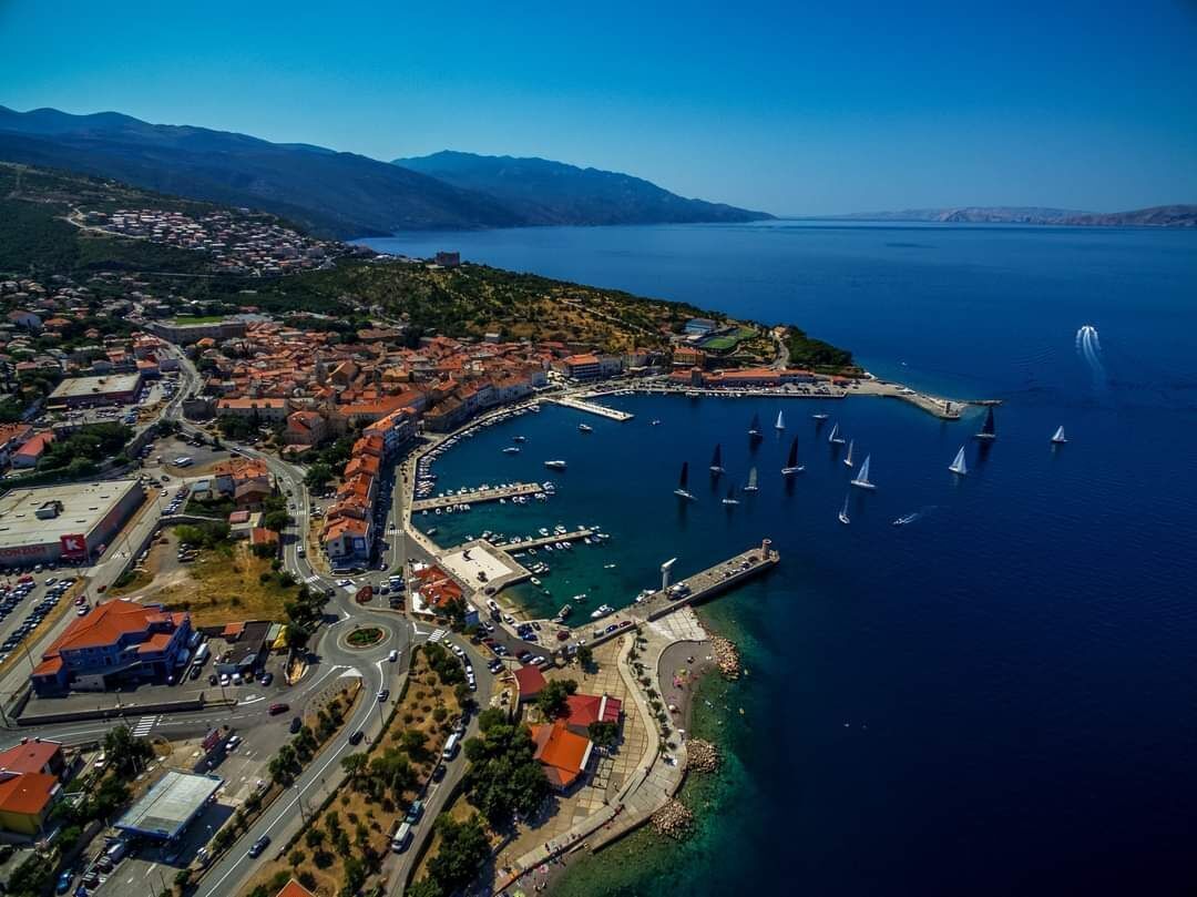 An aerial view of a coastal town with a marina, boats, and a clear blue sea.