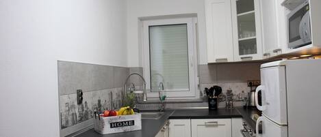 A small kitchen with white cabinets, a black countertop, a window with blinds, and a microwave above the counter.