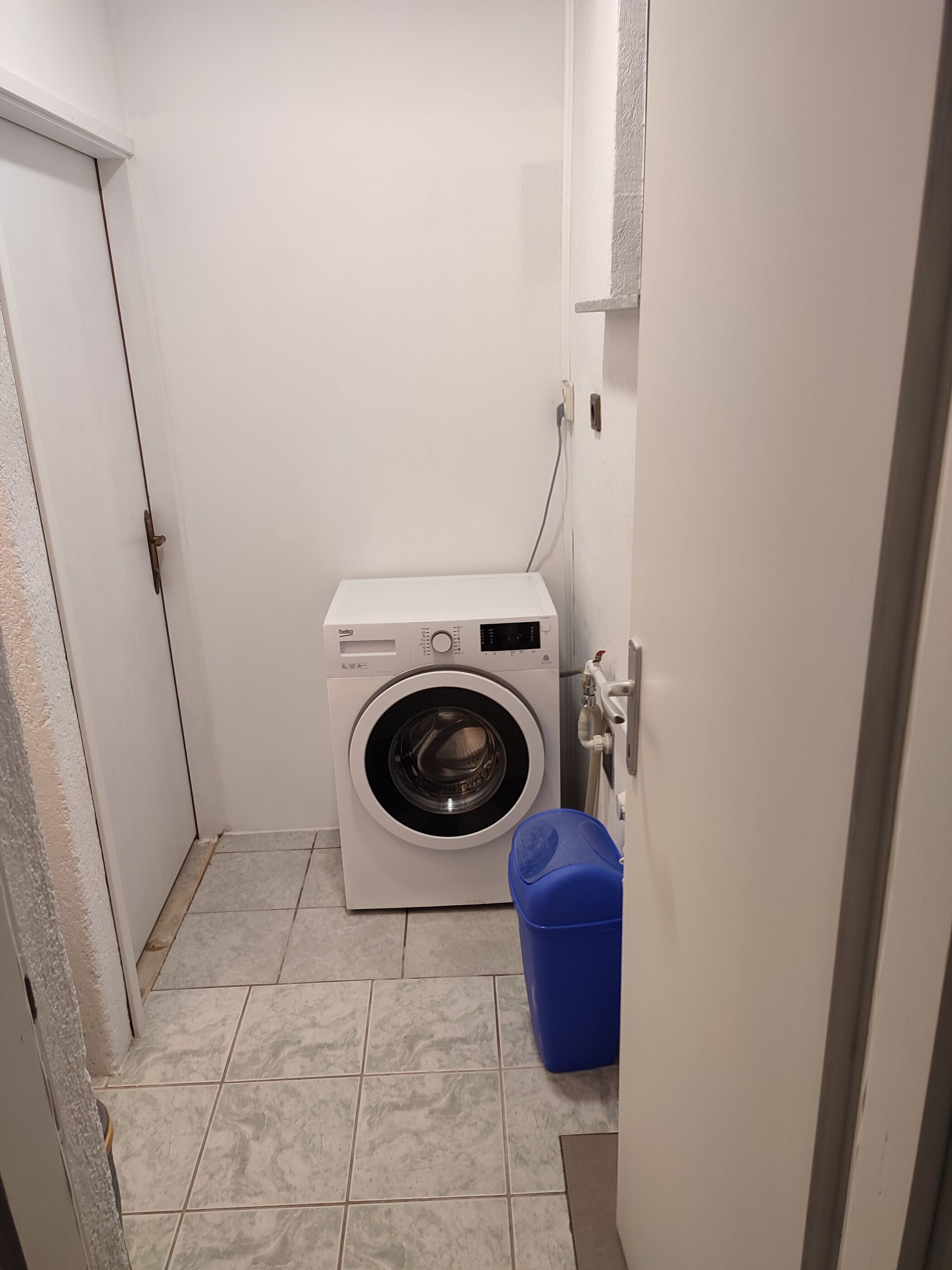 A compact laundry room with a front-loading washing machine, a blue trash bin, and tiled flooring.