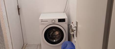 A compact laundry room with a front-loading washing machine, a blue trash bin, and tiled flooring.