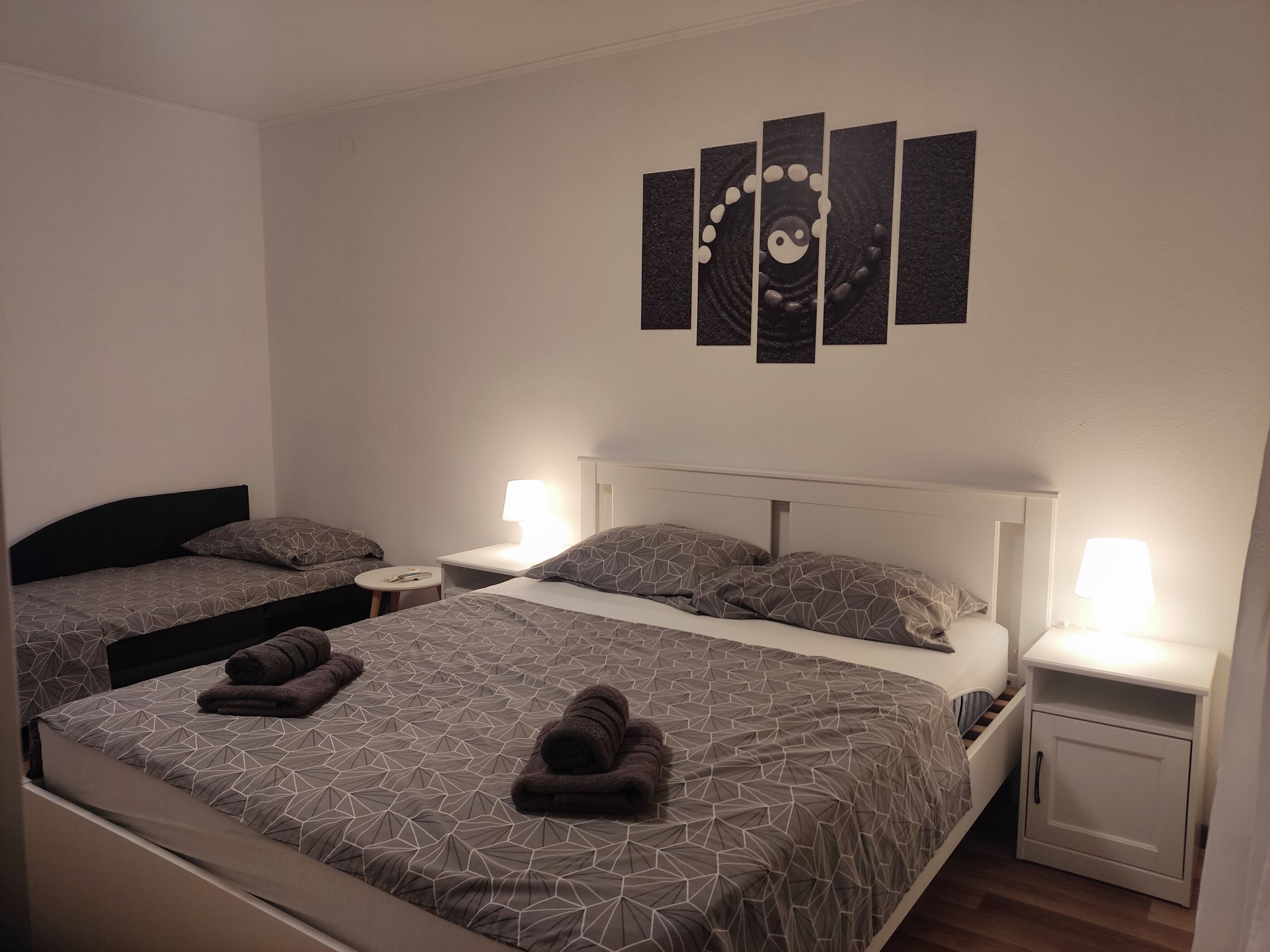 A hotel room with two beds, a bedside table with a lamp, a wall art piece, and a small cabinet.