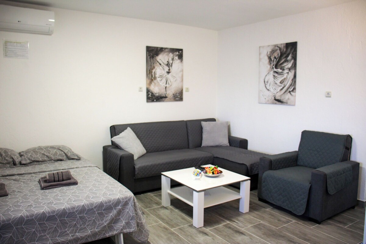 A modern living room with a grey sofa, a smaller grey armchair, and a white coffee table. There are two abstract paintings on the walls and a bed with patterned bedding in the background.