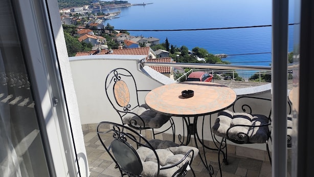 Un balcone con tavolo e sedie, affacciato su una località costiera e il mare.