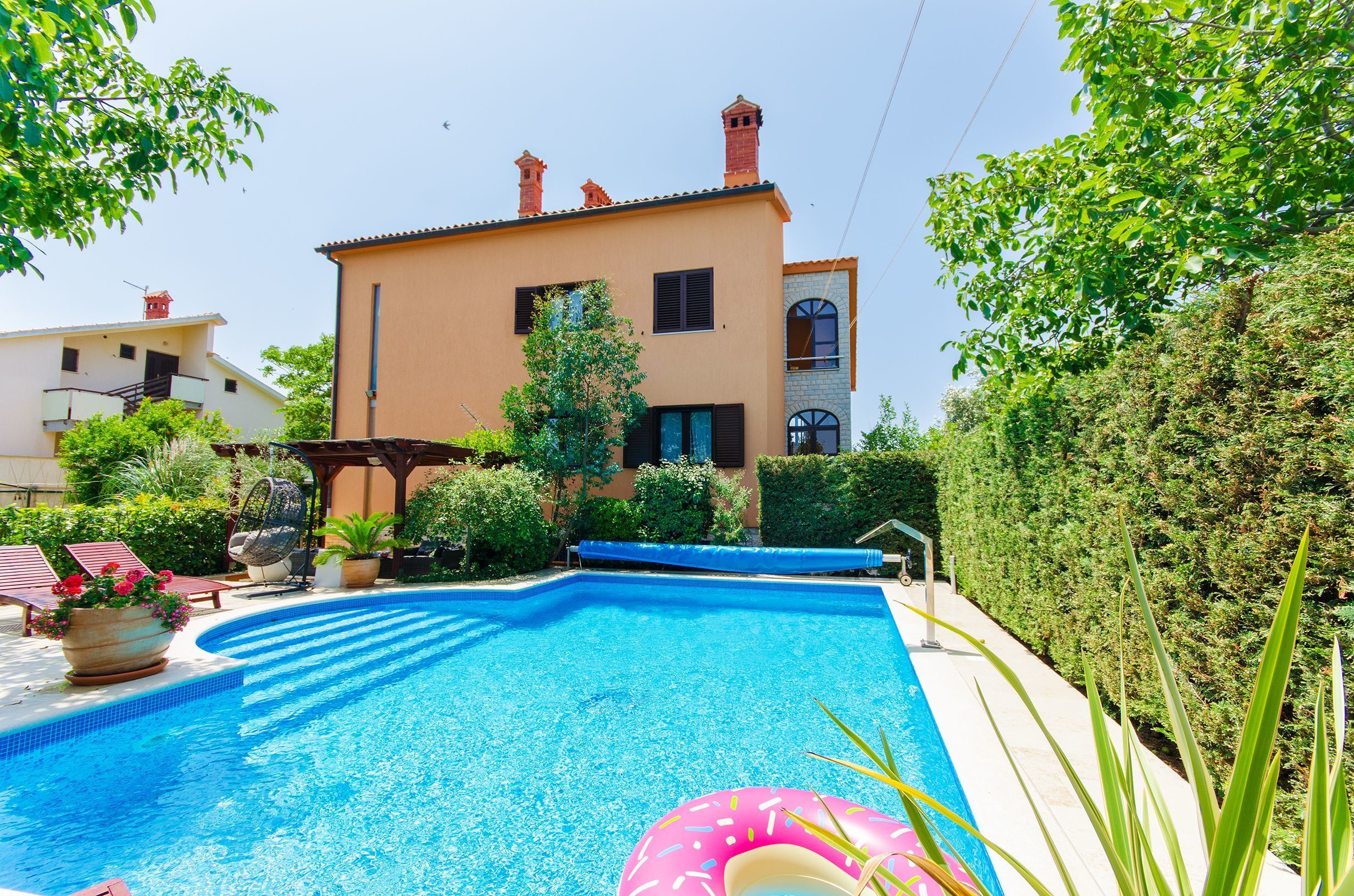 A swimming pool with a pink inflatable ring, surrounded by a garden and a building with a tiled roof.