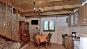 A kitchen with a dining table, chairs, wooden cabinets, a stone wall, and a window with curtains. - House Mariano (Lasici)