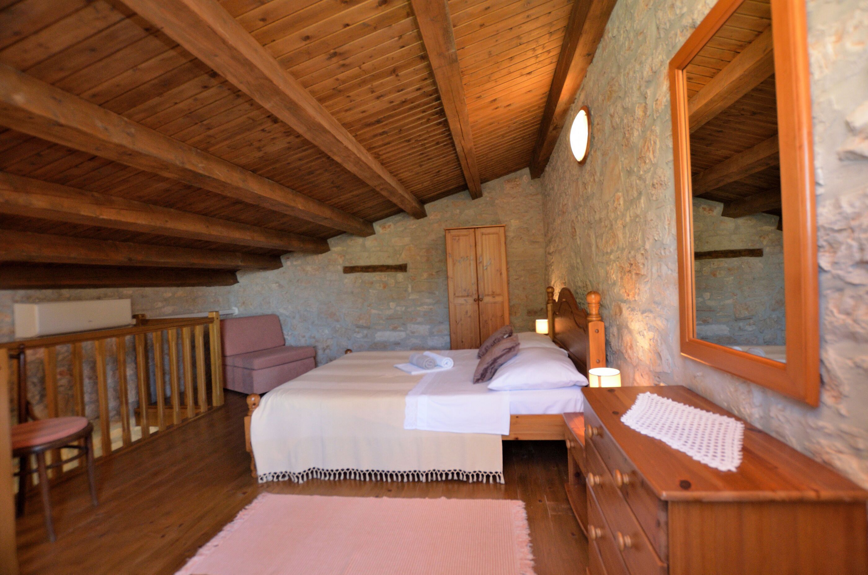 Un dormitorio con techo de madera, paredes de piedra, una cama con sábanas blancas, un aparador de madera y un espejo grande.