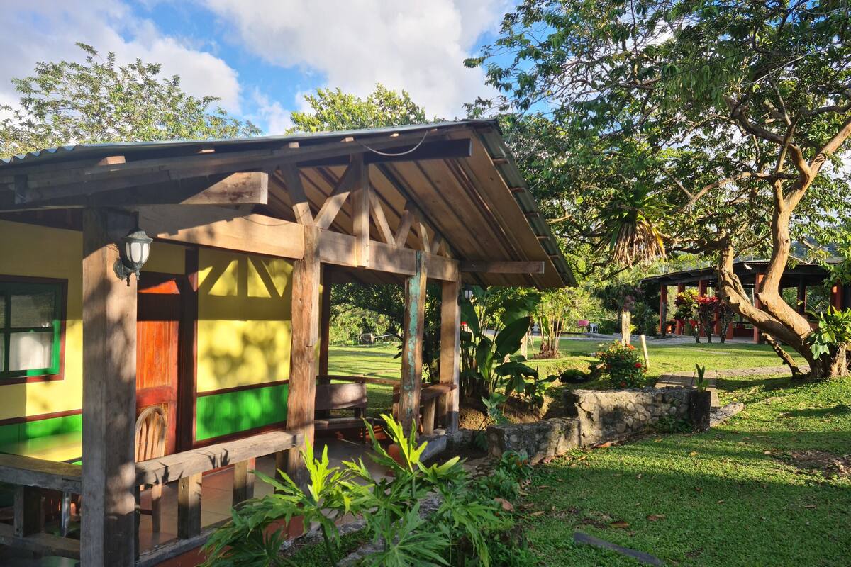 A small, colorful wooden structure with a porch, surrounded by lush greenery and trees.