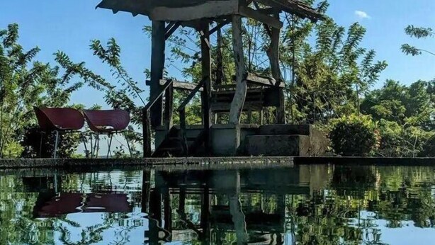 Un quiosco de madera con techo, rodeado por una masa de agua tranquila que refleja árboles y el cielo.