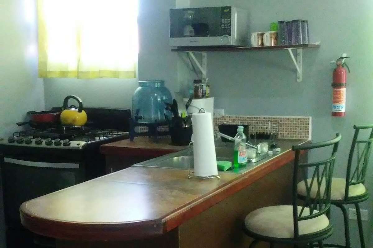 A kitchen with a central island, bar stools, a stove, a kettle, a microwave, and a fire extinguisher.