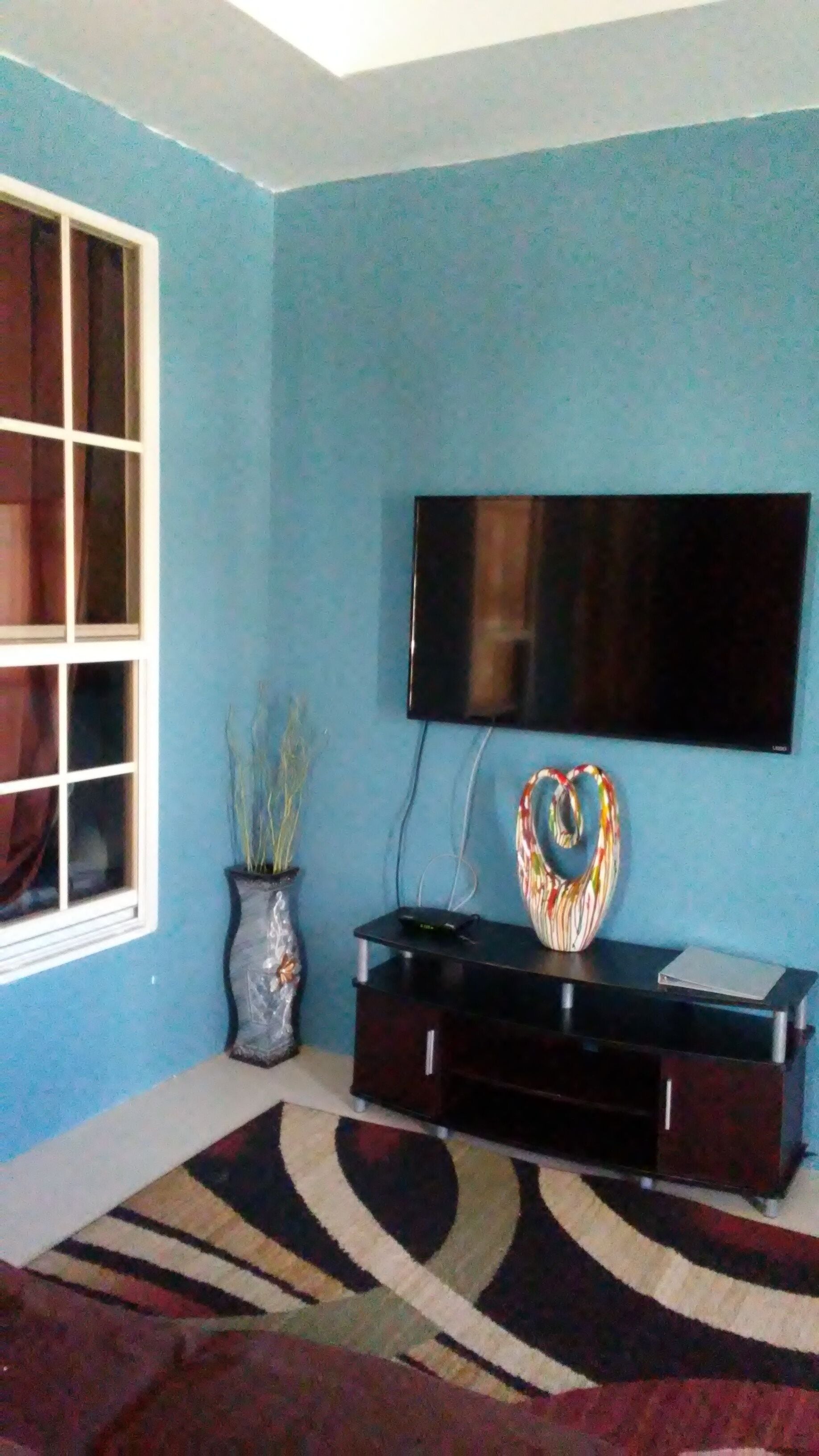 A living room with a blue wall, a flat-screen TV mounted above a dark wooden entertainment unit, a potted plant, and a patterned rug.