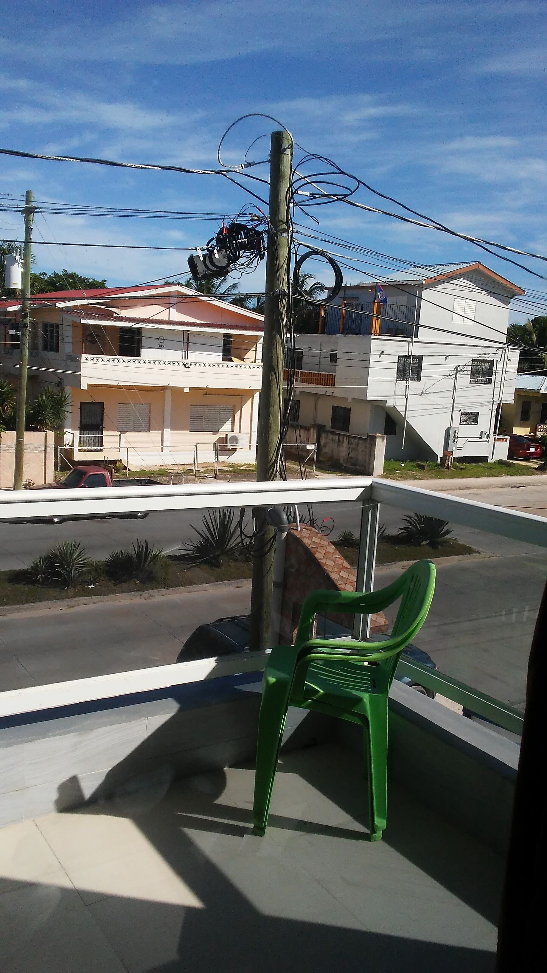 Vista da rua com edifícios, uma cadeira de plástico verde e um poste de eletricidade com fios.