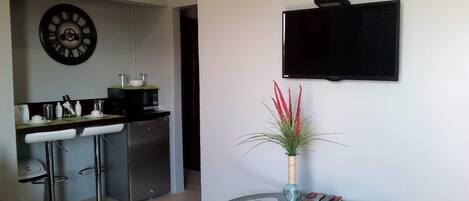 Una cocina moderna con barra, un reloj de pared, un televisor de pantalla plana y una pequeña mesa con un jarrón de flores rojas.