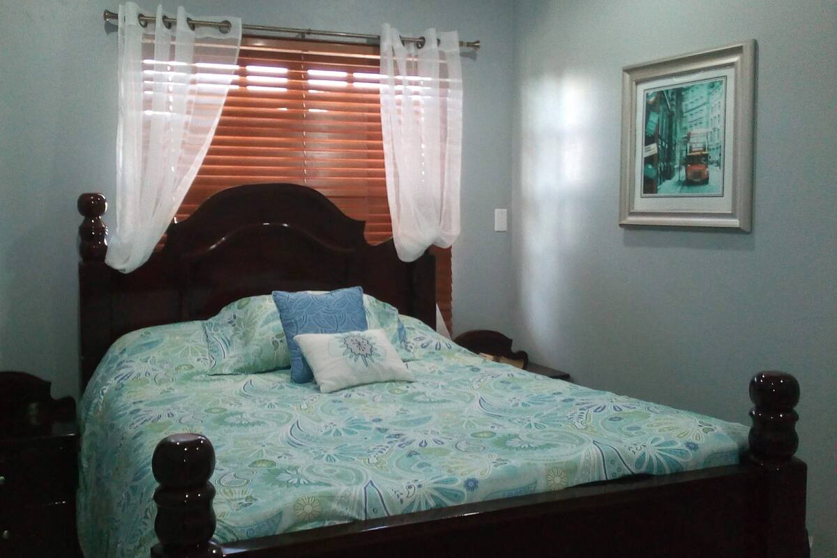 A bedroom with a bed, a nightstand, a window with blinds, and a framed picture on the wall.