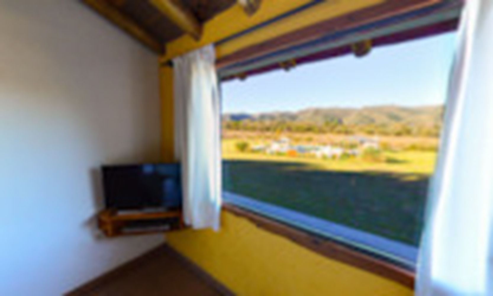 Ein Zimmer mit Fernseher auf einem Holzständer, einem Fenster mit Blick auf eine grüne Landschaft und Gebäude sowie einer gelben Wand.