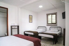 A room with two single beds, a large bed, a mirror, a shelf with towels, a framed picture, and a window with blinds. - Finca Hotel Los Guayacanes (San José)