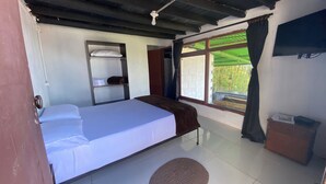 Una habitación con cama, estante con ropa doblada, ventana con cortinas y una pequeña mesa con un bolso encima.