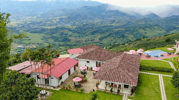 Un hotel con varios edificios de techo rojo, áreas de estar al aire libre con sombrillas y vistas a colinas verdes y exuberantes al fondo.