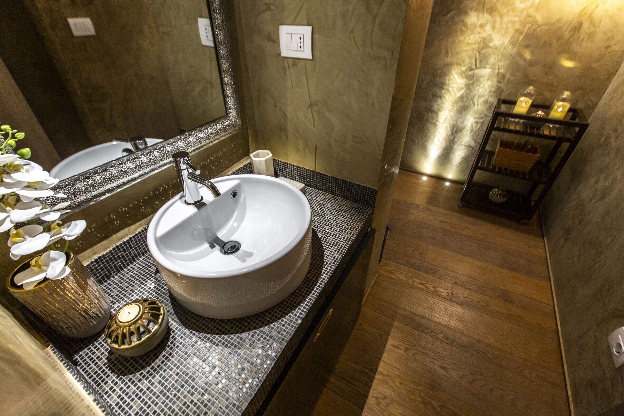 A bathroom with a round sink, a mirror, a small shelf with candles, and a wooden floor.