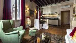 Un salon avec un fauteuil vert, un canapé blanc, un petit pouf et un tapis à motifs. On y trouve également un coin repas avec une table et des chaises, et une cuisine à l’arrière-plan.