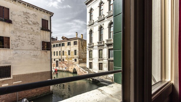 Une vue sur un canal, depuis une fenêtre donnant sur un balcon, avec en arrière-plan des bâtiments historiques et un pont.