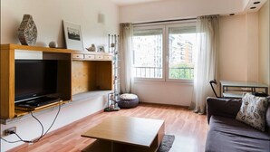 Un salon moderne doté d’un téléviseur à écran plat, de meubles en bois, d’une table basse et offrant une vue sur les bâtiments grâce à de grandes fenêtres.