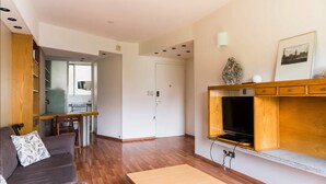 Un salon moderne avec un canapé, une table basse en bois, un téléviseur fixé sur un meuble en bois, et un coin cuisine à l’arrière-plan.