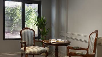 Ein Zimmer mit zwei Polstersesseln, einem kleinen runden Tisch, einer Topfpflanze und einem roten Teppich mit Muster auf Fliesenboden.
