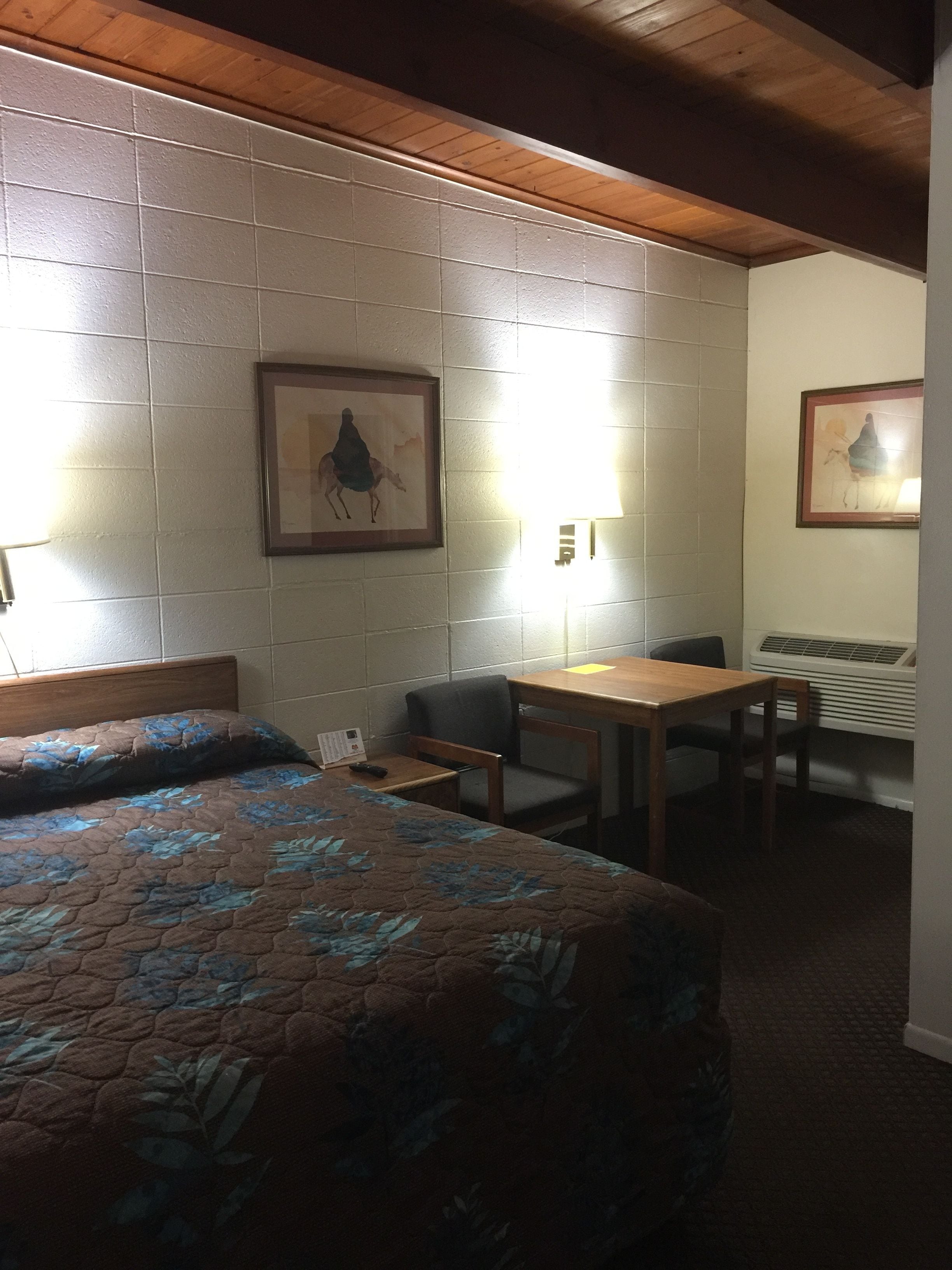 A hotel room with a bed, a small table, a chair, and a desk. There are two framed pictures on the wall.