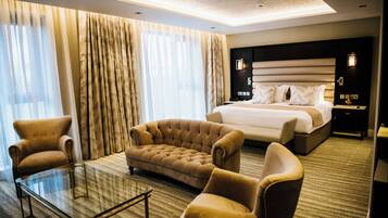 A hotel room with a large bed, two armchairs, a glass coffee table, and a view of the outdoors through sheer curtains.