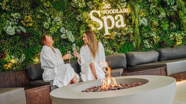 Two people in white robes sitting on a grey sofa in front of a wall with a 'The Woodland Spa' sign.