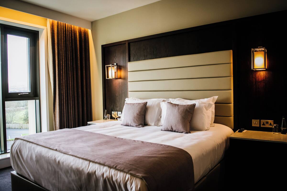 Habitación de hotel con una cama grande, dos lámparas de noche, una ventana con cortinas y una mesita de noche con un teléfono y un vaso.