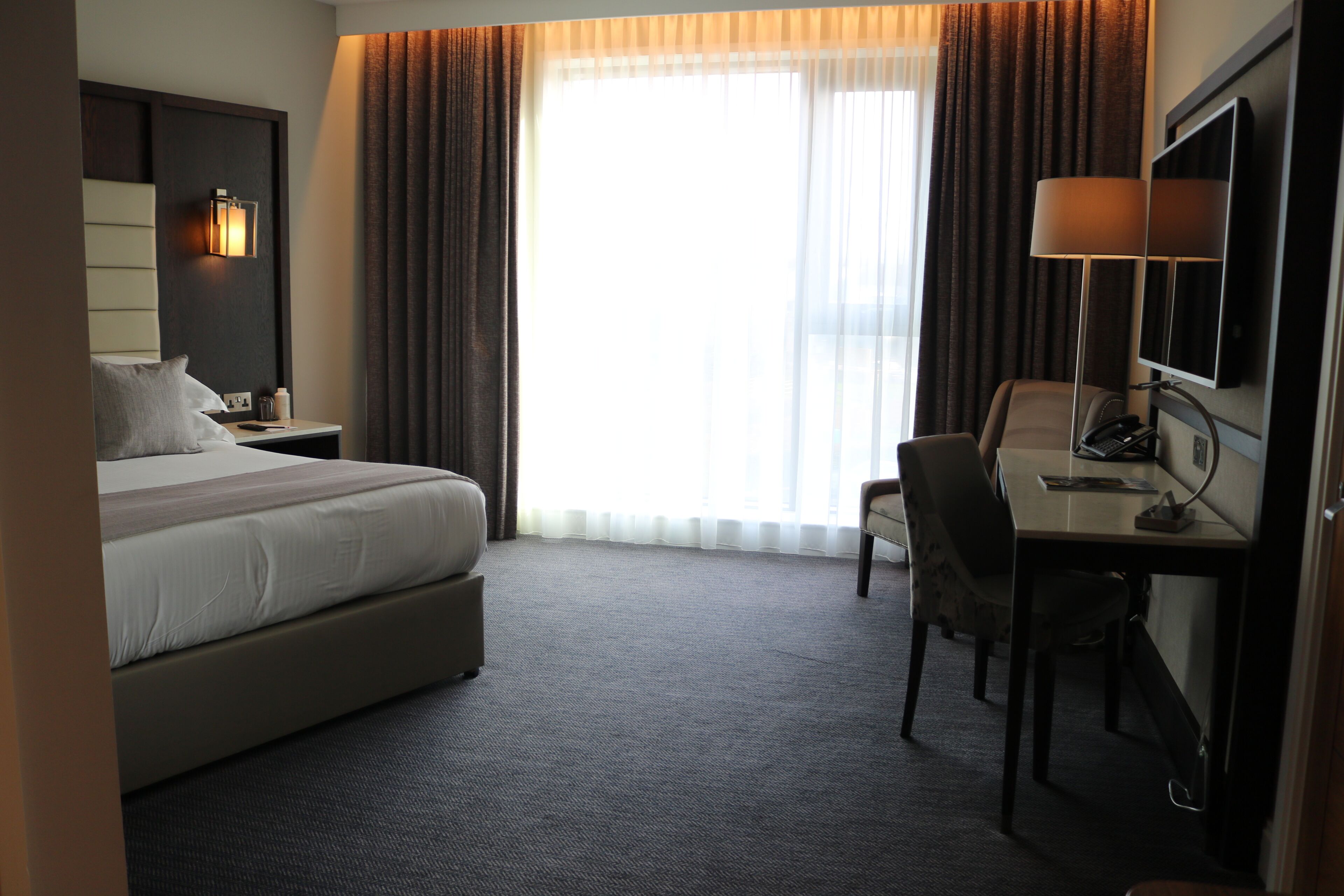 A hotel room with a bed, desk, chair, and a large window with curtains.