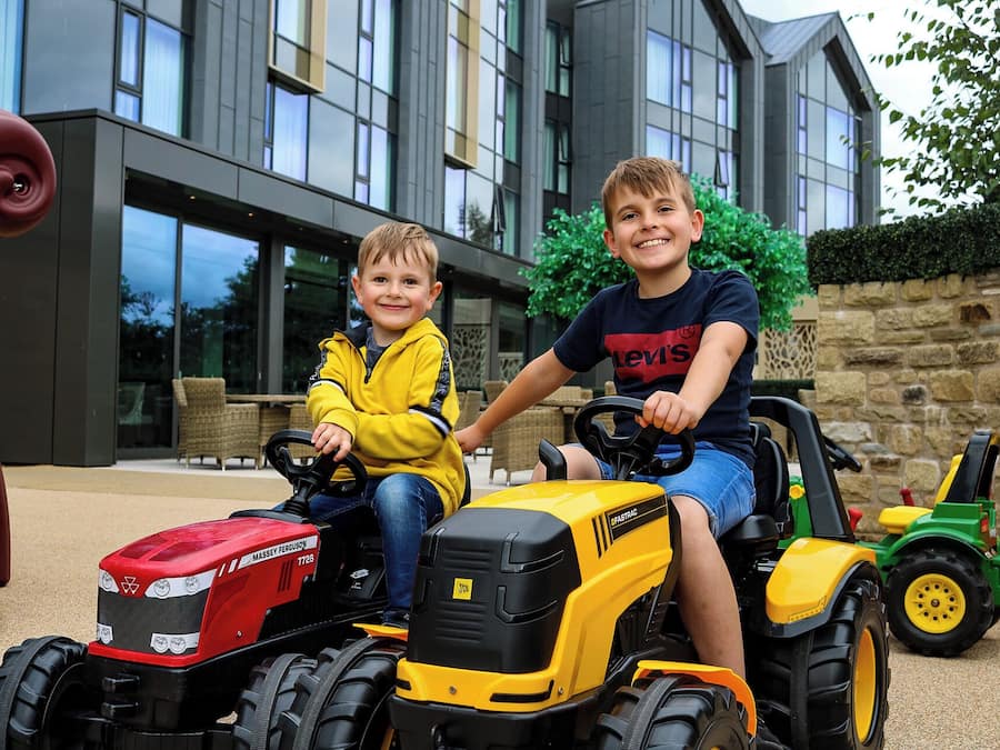 Two children on a tractor in front of a modern building with large windows and a stone wall.