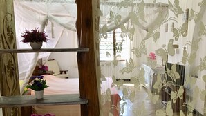 Une pièce avec un plafond en bois, un lit à baldaquin blanc et une fenêtre donnant sur l’extérieur. Des plantes en pot sont posées sur le rebord de la fenêtre, et un rideau décoratif orné d’un motif floral complète l’ensemble.