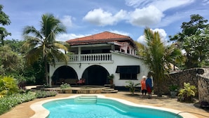 Une piscine à bords incurvés, un bâtiment blanc surmonté d’un toit recouvert de tuiles rouges et des palmiers.