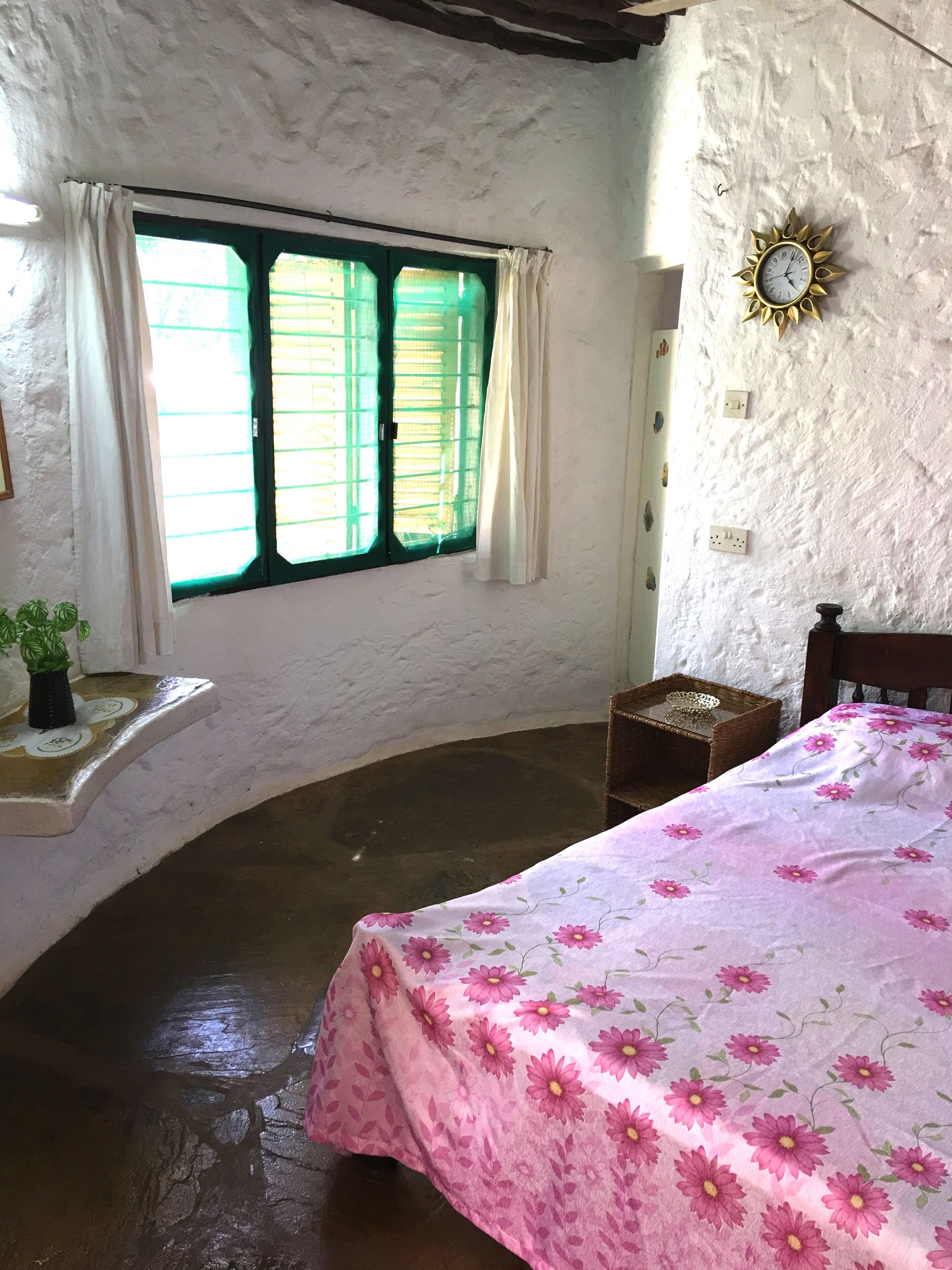 A bedroom with a bed, a window with curtains, a wall clock, and a small shelf with a plant.