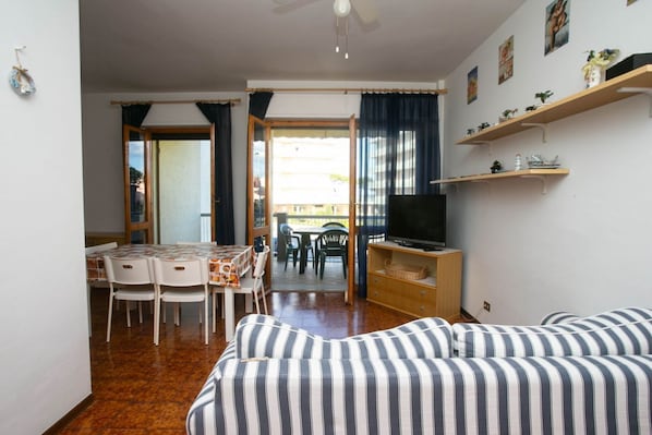 A furnished room with a sofa, a TV on a wooden stand, a dining area with chairs and a table, and a balcony with a view.