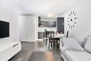Una sala moderna con una televisión de pantalla plana, un comedor con mesa y sillas, y una cocina al fondo.