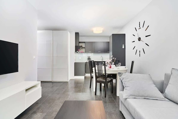 Una sala moderna con una televisión de pantalla plana, un comedor con mesa y sillas, y una cocina al fondo.
